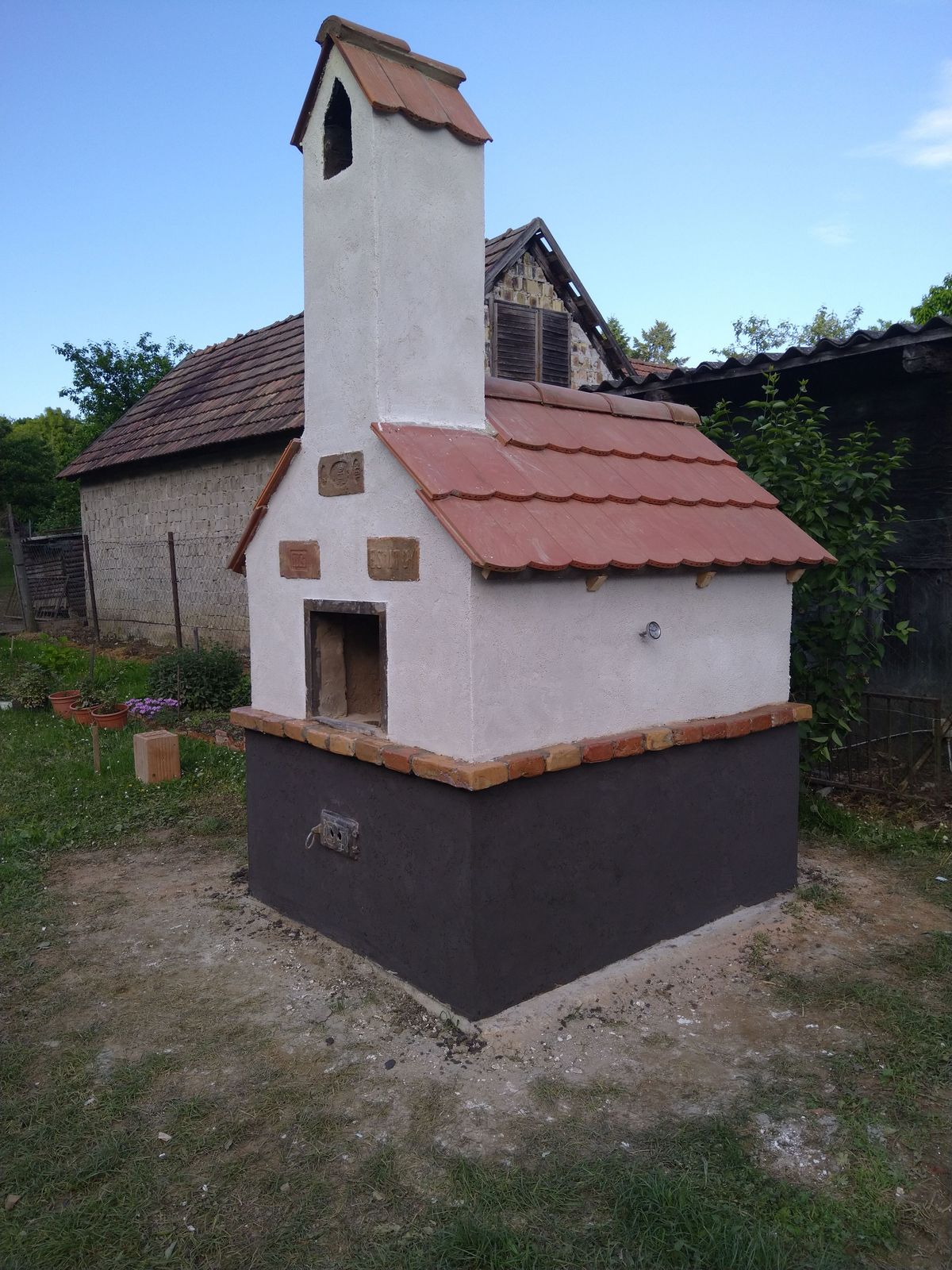 Fehér felületű kerti kemence piros cseréptetővel, magas kéménnyel és sötét lábazattal.