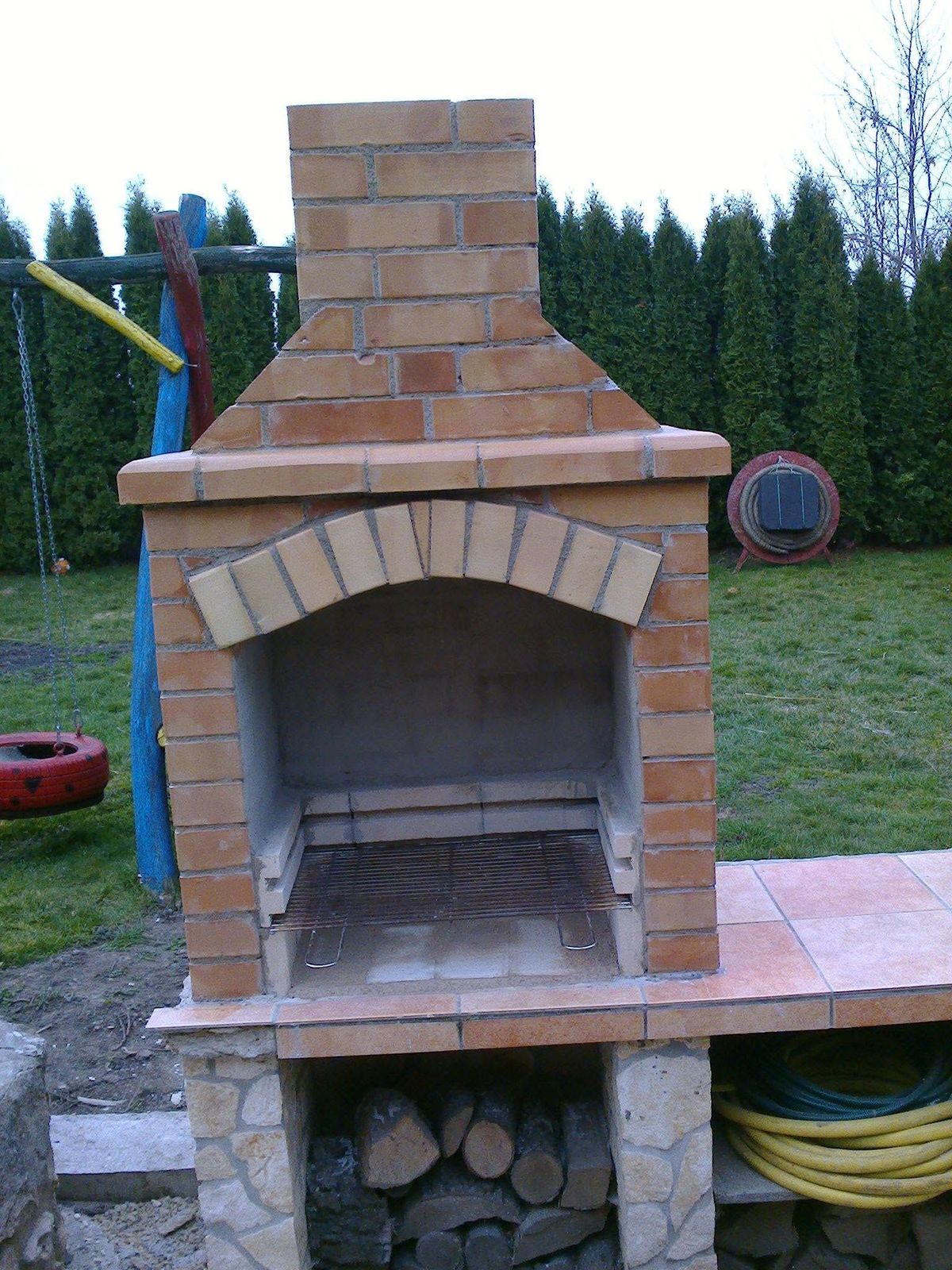 Nyitott kerti grillsütő íves téglakerettel, kéménnyel, oldalsó munkafelülettel és alsó tárolóval.